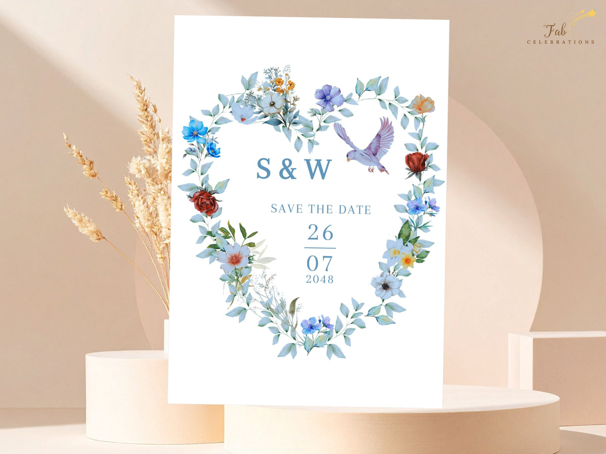 Save the date wedding announcement featuring watercolor-inspired blue flowers and fresh green foliage arranged in a heart-shaped wreath with two doves framing the couple's initials and date.