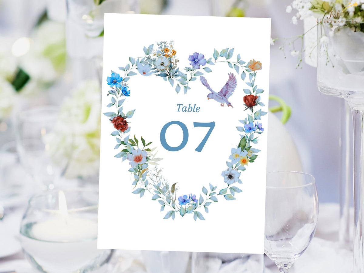 Wedding table number card featuring a large blue table number framed by a watercolor-inspired floral heart wreath with dusty blue blooms, greenery and two delicate doves for coordinated reception decor.