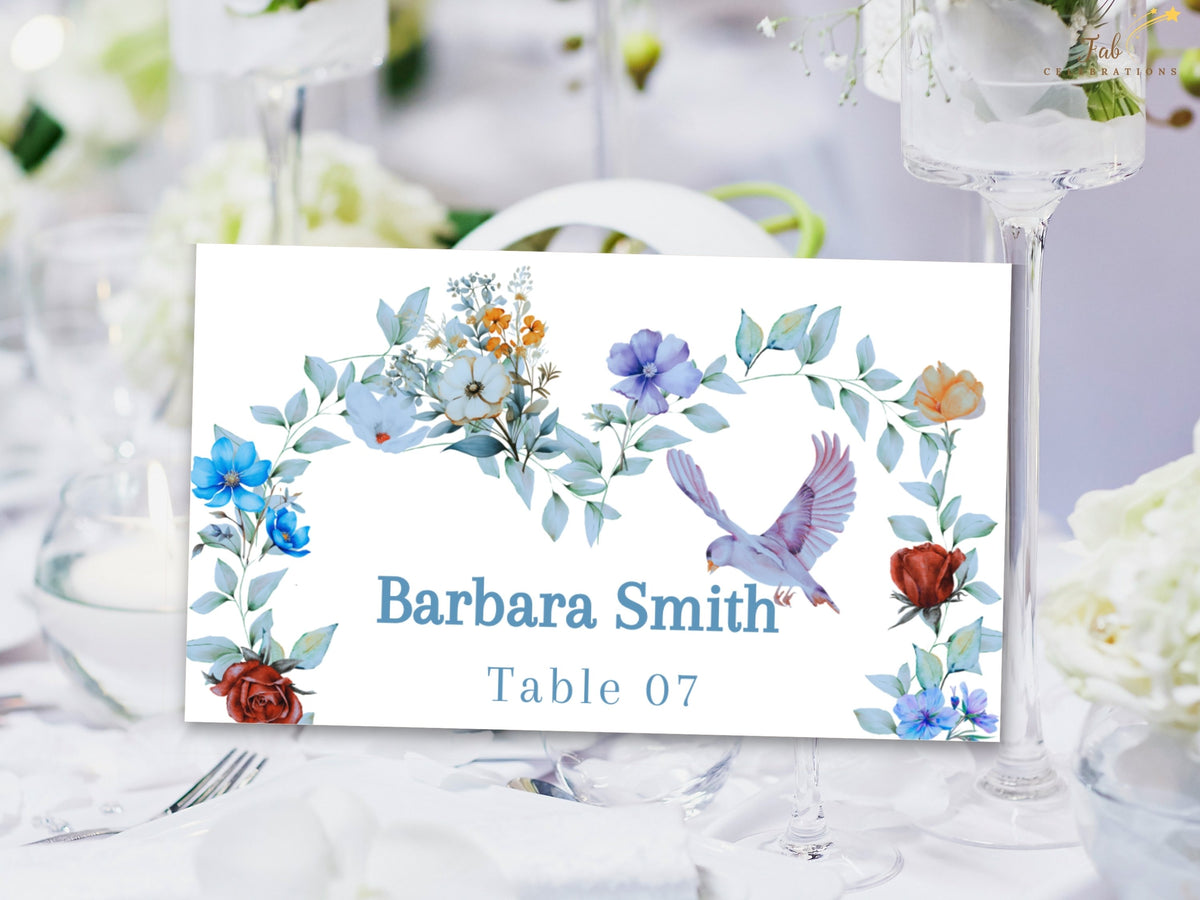 Wedding place card featuring a guest name and table assignment framed by watercolor-inspired dusty blue flowers, greenery and a graceful dove, designed to coordinate with the full wedding stationery collection.