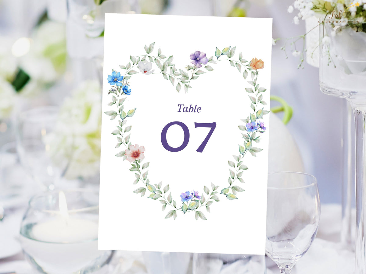 Wedding table number card featuring a watercolor-inspired heart-shaped wreath of airy green foliage with blue, purple, white and peach flowers surrounding a large elegant purple table number.