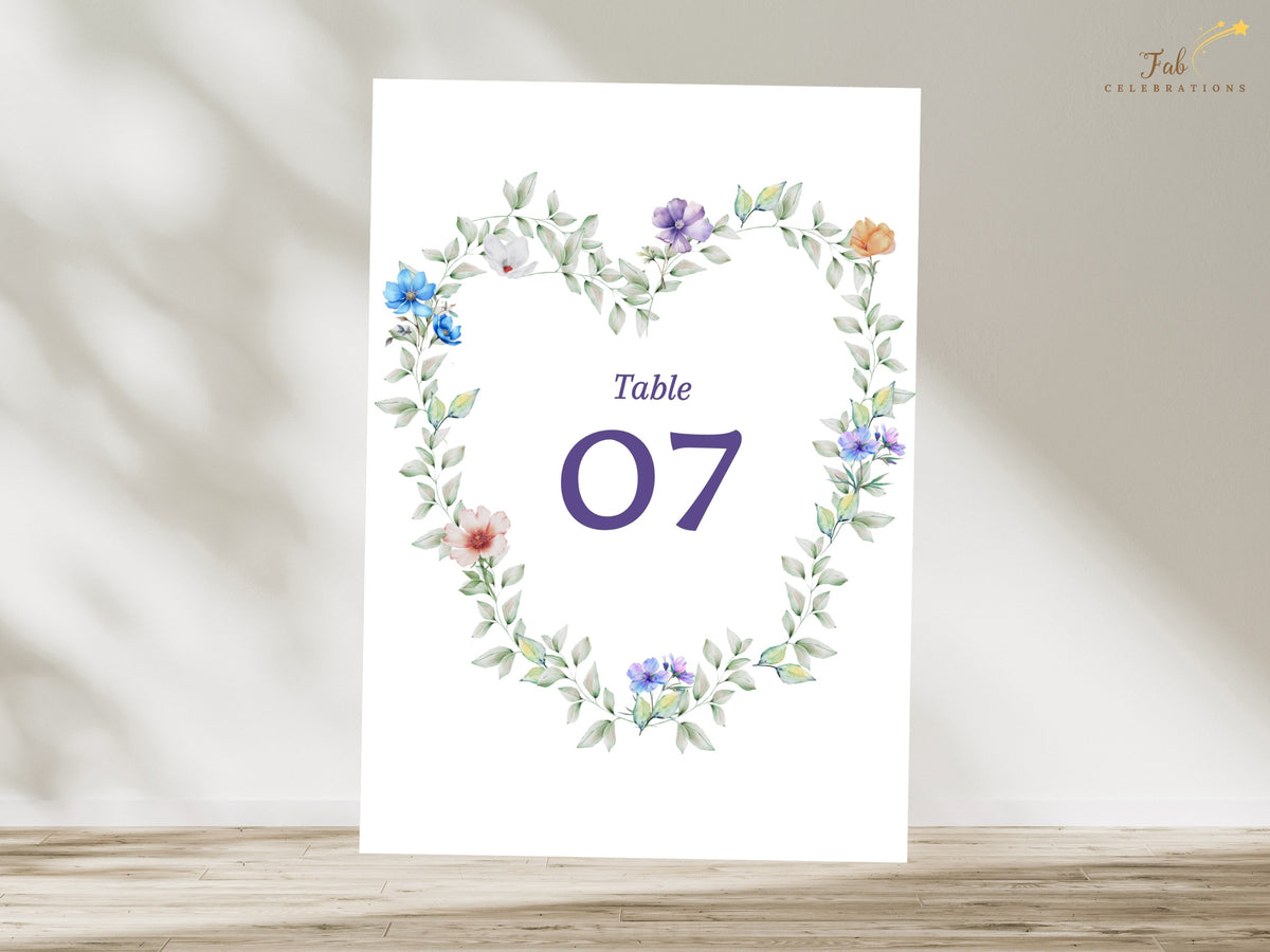 Wedding reception table number featuring a watercolor-inspired botanical wreath of delicate blue, purple, white, peach and pink flowers with airy greenery around an elegant purple number.