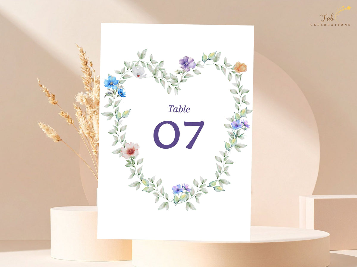 Wedding table card with a large elegant purple number centered inside a watercolor-inspired floral wreath of soft blue, purple, white, peach and pink flowers with delicate green foliage.