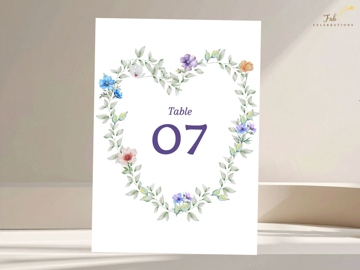 Wedding table number card decorated with watercolor-inspired soft pastel flowers and airy green foliage arranged in an elegant wreath around a prominent purple reception table number.