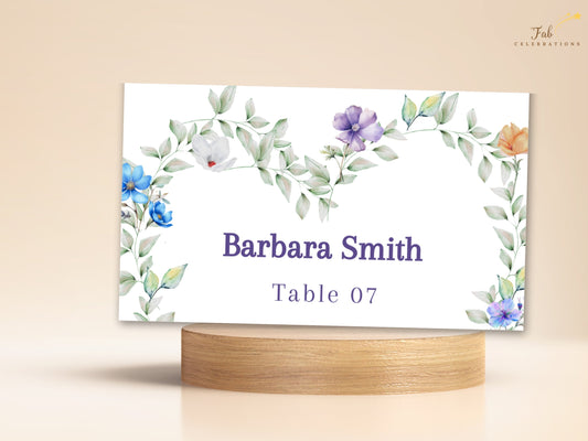 Pastel Wedding Name Card featuring an editable guest name and table number framed by watercolor-inspired airy green foliage and delicate blue, purple, white, peach and pink flowers for a coordinated wedding place setting.