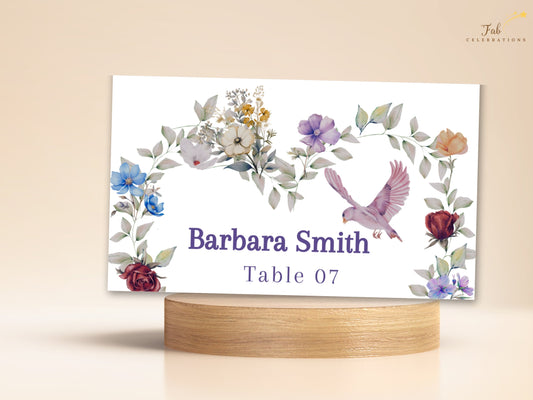 Purple floral wedding place card showing Barbara Smith and Table 07 inside a watercolor-inspired heart of violet, blue, blush and burgundy flowers, with a pink dove and soft green foliage.
