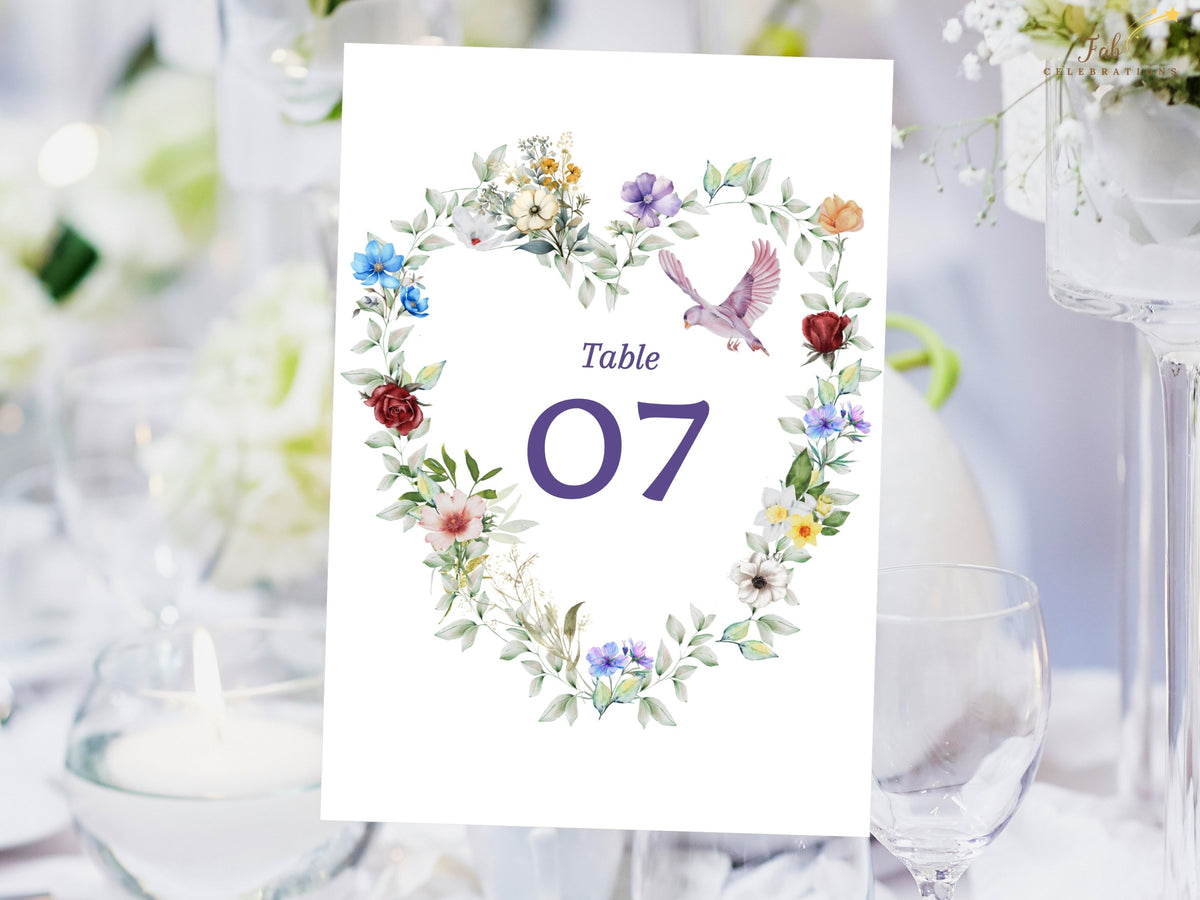 Wedding table number card featuring a watercolor-inspired floral heart with multicolored blossoms, soft leafy greenery and a flying dove surrounding a large elegant table number.
