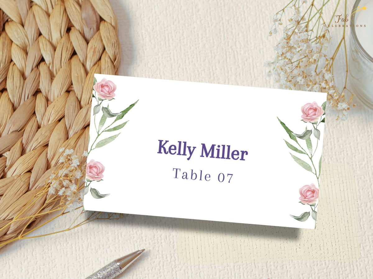 Pink floral seating card decorated with watercolor-inspired blush roses and green foliage, personalized with an elegant guest name and clearly displayed reception table number.